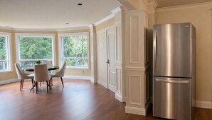 A modern kitchen with a stainless steel refrigerator, wooden flooring, and large windows offering a view of greenery.