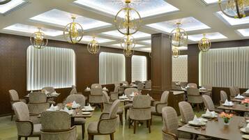 A spacious dining area with multiple tables and chairs, ornate chandeliers, and large windows with blinds.
