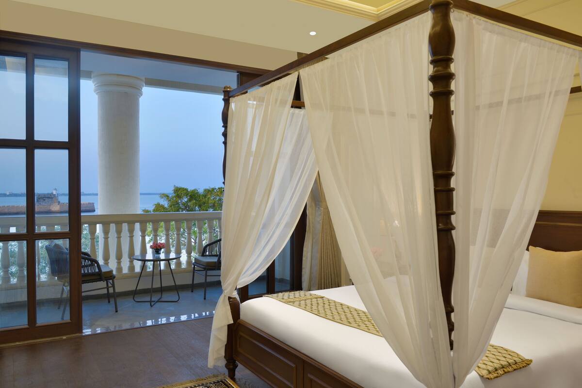 A four-poster bed with sheer curtains, a balcony with a table and chairs, and a view of the sea.