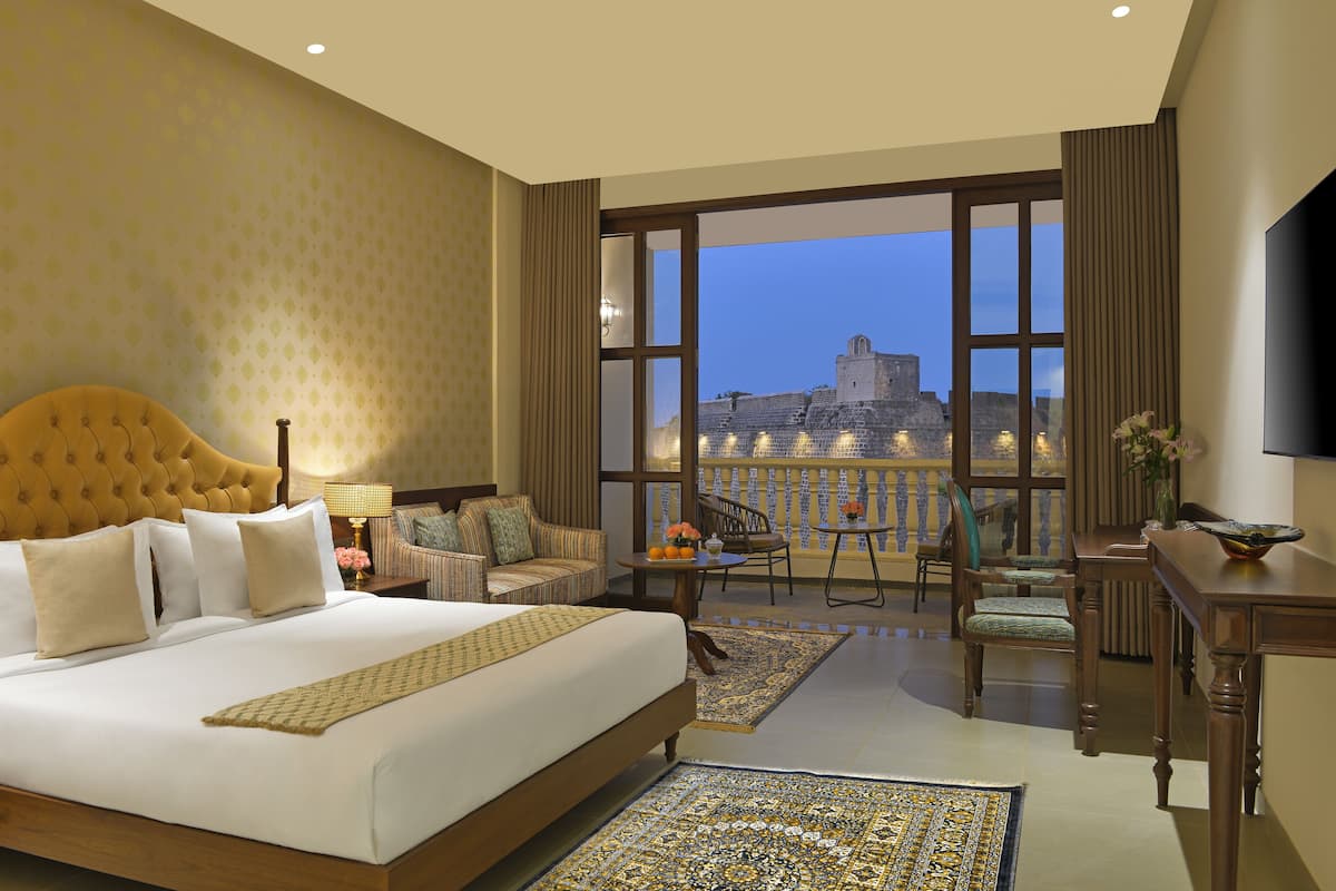 A hotel room with a large bed, a balcony with a view of a historic building, a dining area with chairs and a table, and a TV on the wall.
