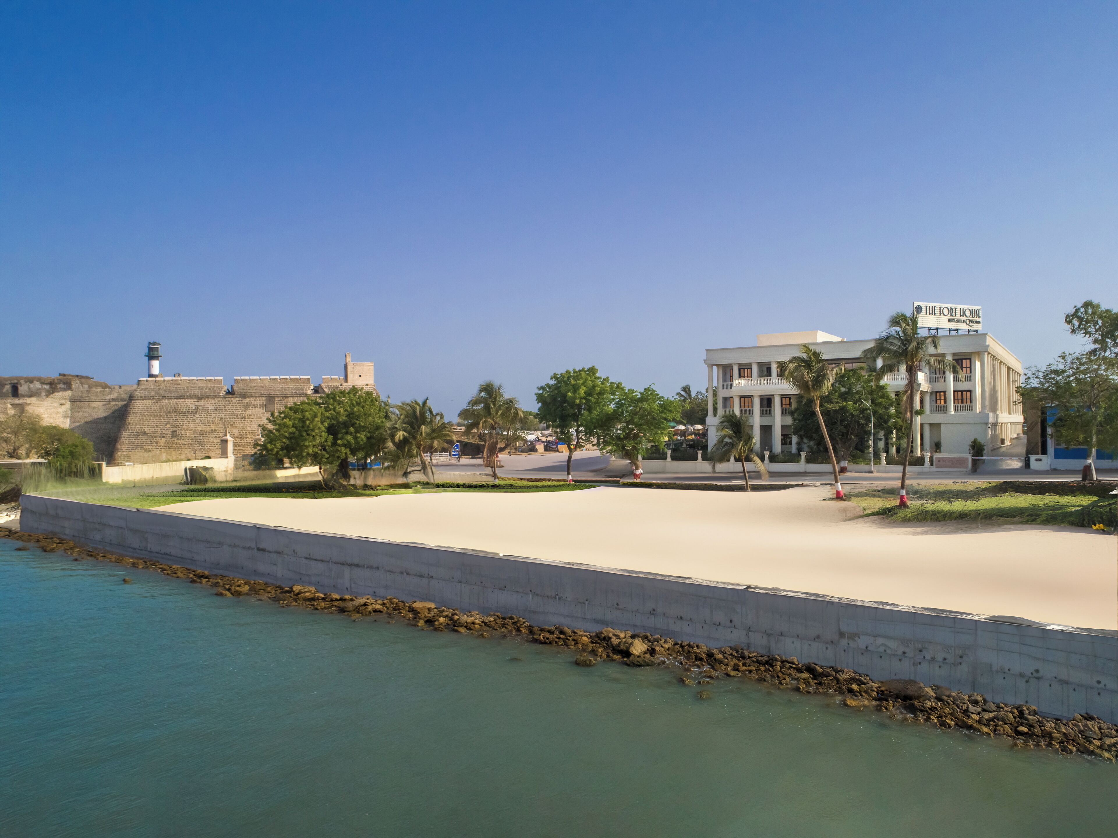 Un hôtel situé sur la côte, doté d’une plage de sable, d’une structure fortifiée et d’une promenade bordée de palmiers.
