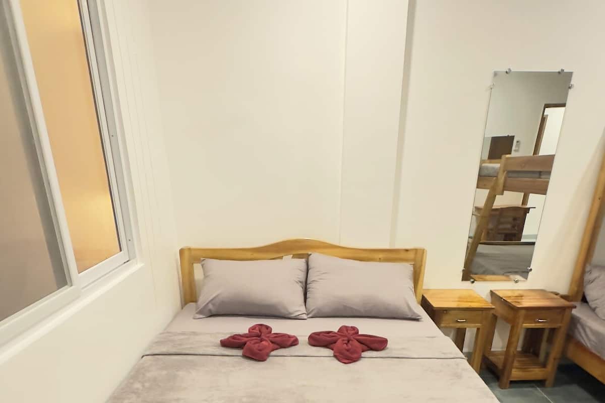 A single bed with a wooden frame and a grey bedspread, two pillows, and two red towels folded in a cross shape. There are two wooden nightstands with drawers, a mirror on the wall, and a window with blinds.