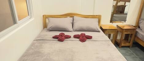 A single bed with a wooden frame and a grey bedspread, two pillows, and two red towels folded in a cross shape. There are two wooden nightstands with drawers, a mirror on the wall, and a window with blinds.