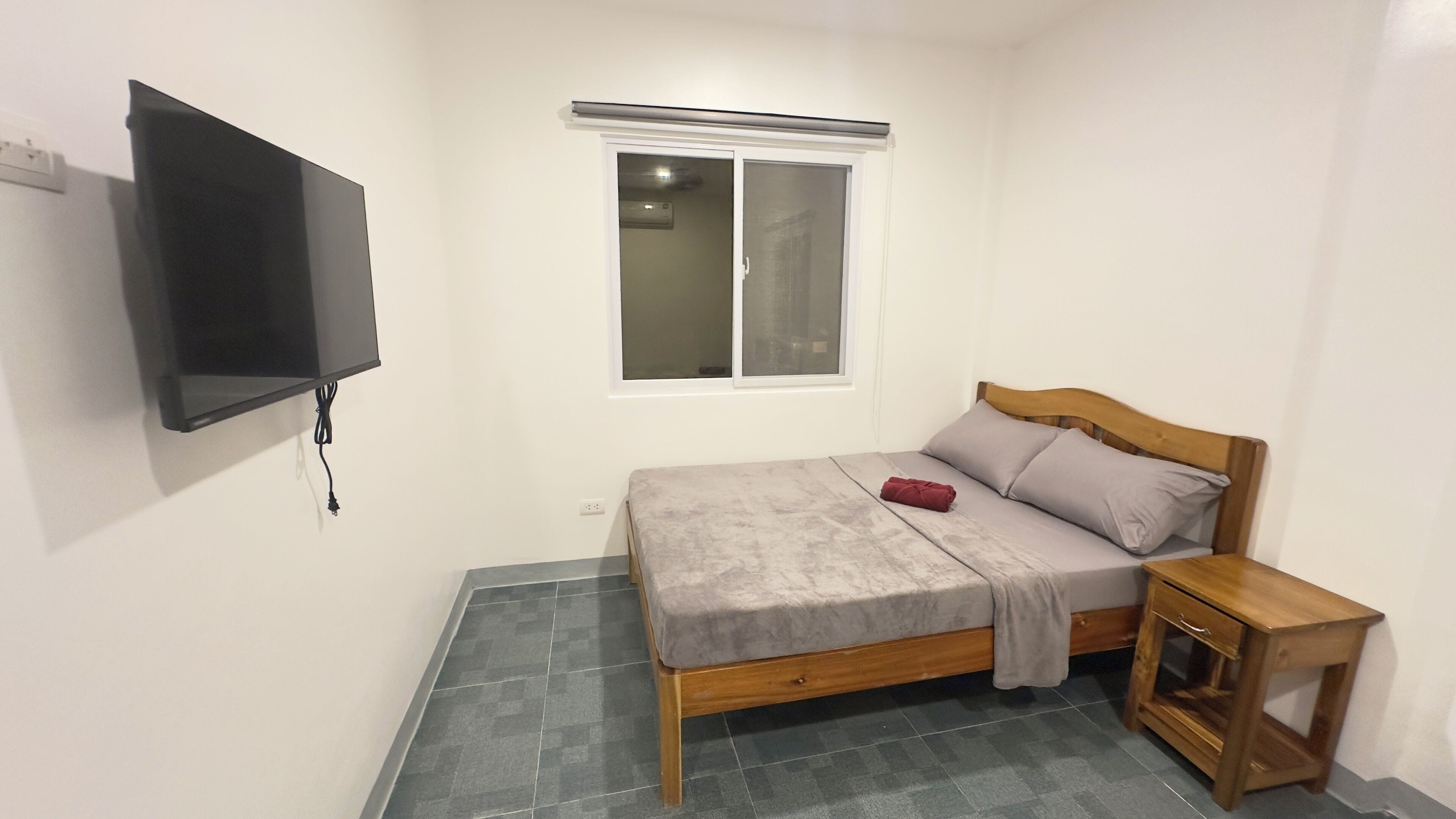 Un dormitorio pequeño y ordenado con una cama, una mesita de noche de madera, un televisor instalado en la pared y una ventana con persianas.