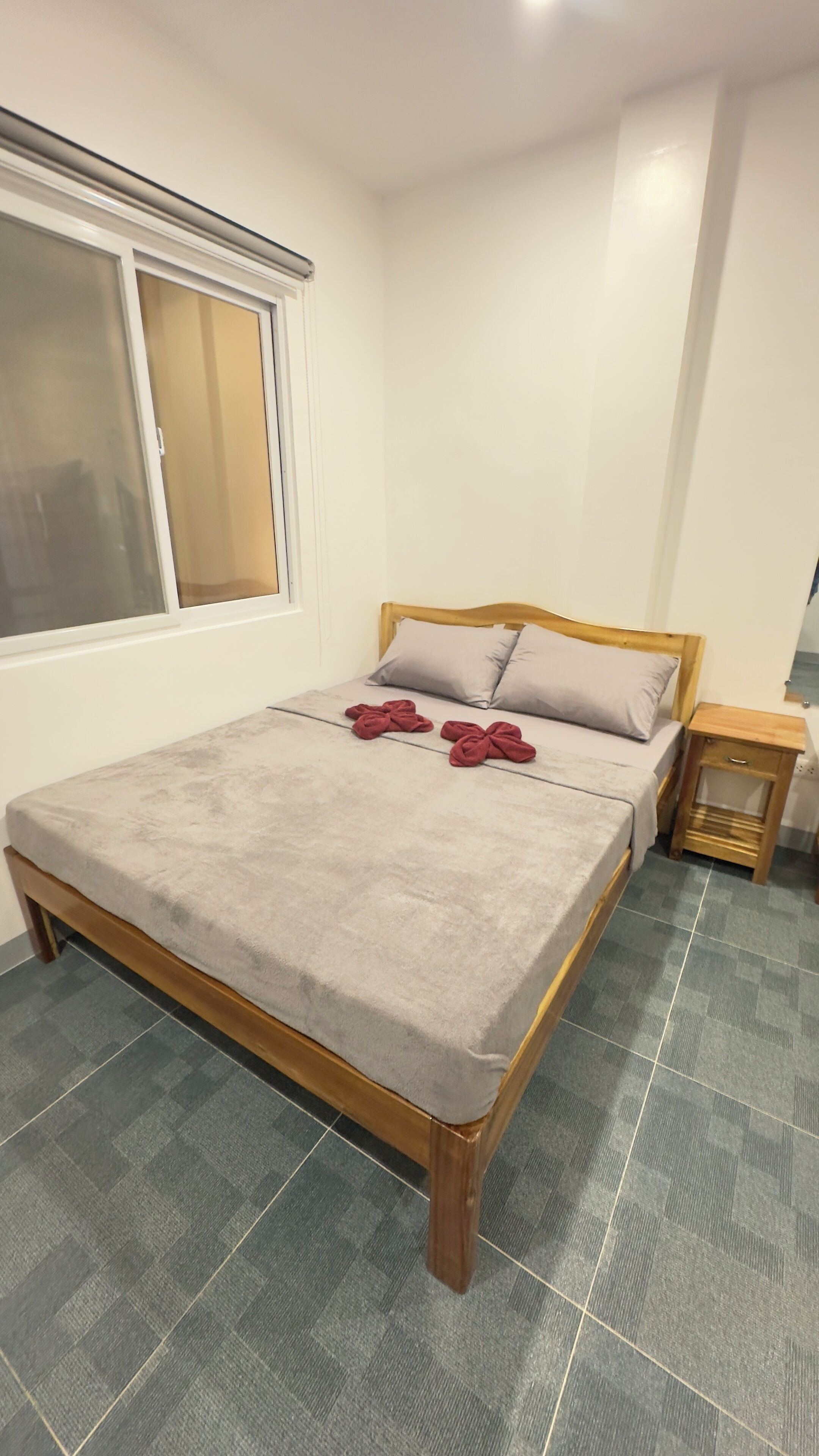 A neatly made bed with a grey blanket and two red towels, a wooden headboard, a small wooden nightstand, and a window with blinds.