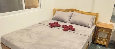 A neatly made bed with a grey blanket and two red towels, a wooden headboard, a small wooden nightstand, and a window with blinds.