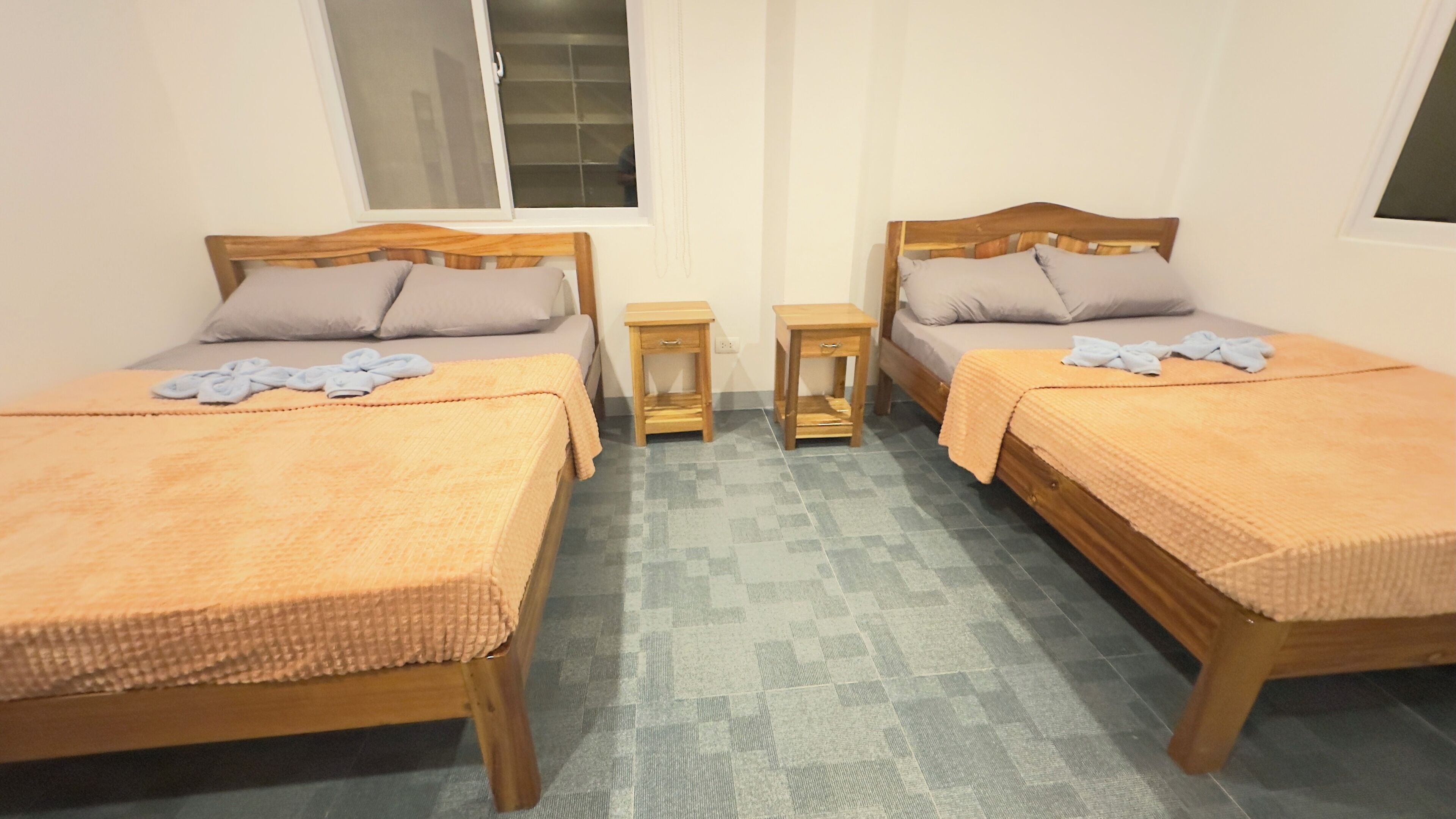 A room with two wooden beds, each with a matching headboard, a small wooden nightstand, and a window with blinds.