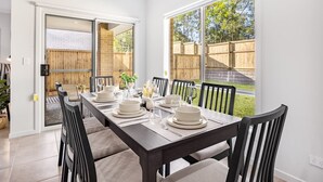 Comedor con una mesa larga dispuesta para seis personas, rodeada de sillas, y vistas a una valla de madera y vegetación al exterior.