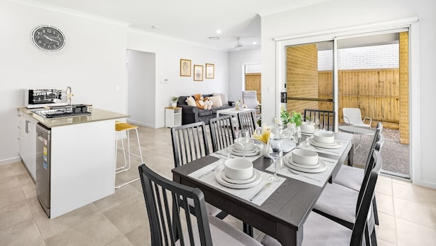 Una cocina moderna con una isla central, comedor con mesa dispuesta, y vista a una valla de madera a través de una puerta de vidrio.