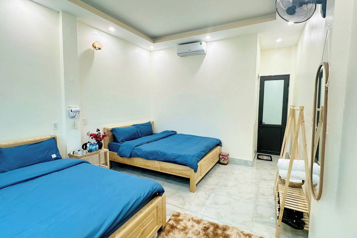 A hotel room with two single beds, a wooden wardrobe, a mirror, a fan, and a small table with a vase and water bottles.