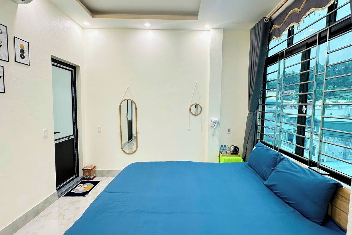 A bedroom with a large bed, a window with a view of buildings, a mirror, and a door.