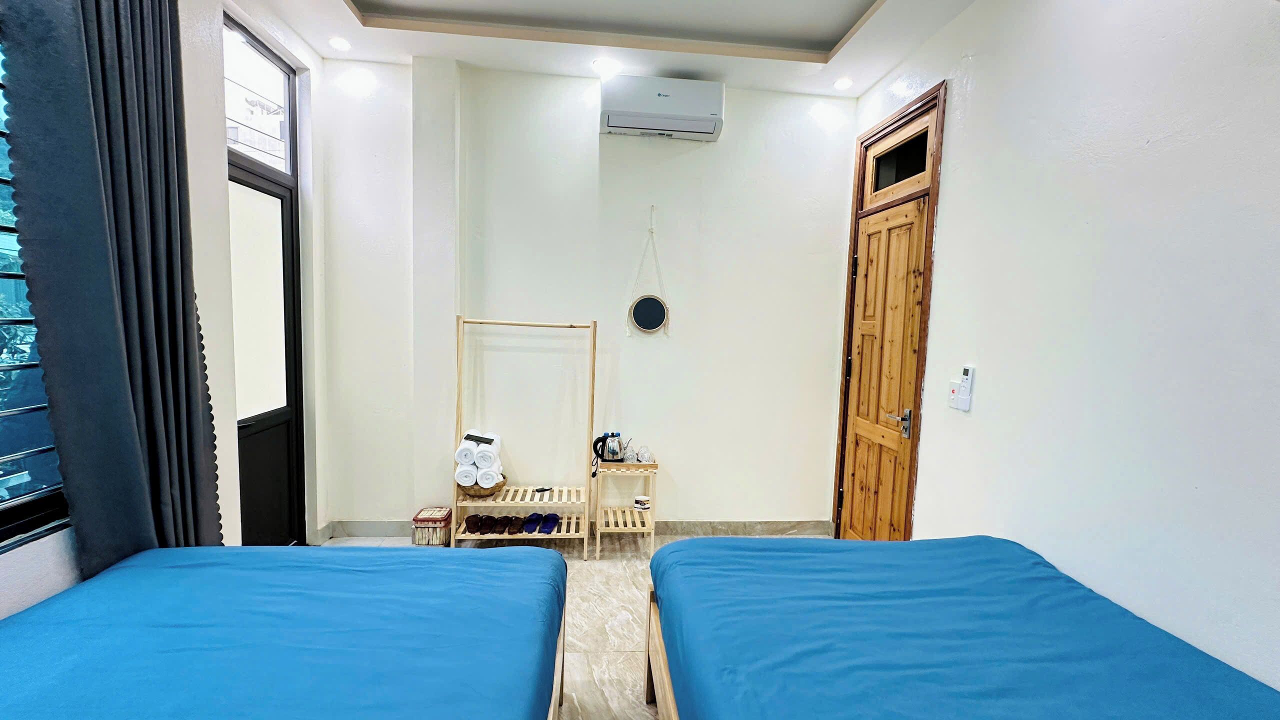 Una habitación con dos camas, una puerta de madera, una mesita con artículos de tocador y una ventana con cortinas.