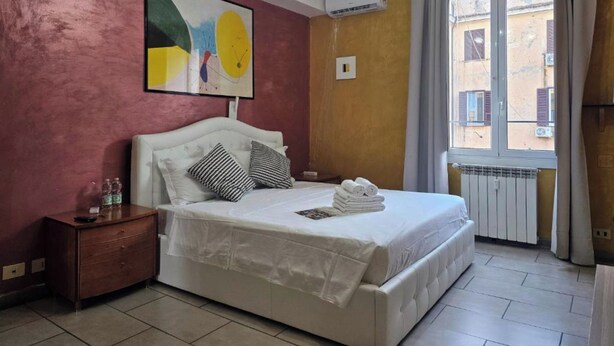 A bedroom with a bed, a nightstand, a painting, an air conditioning unit, and a window with curtains. - Giovanni Giolitti 177 Termini (Rome)