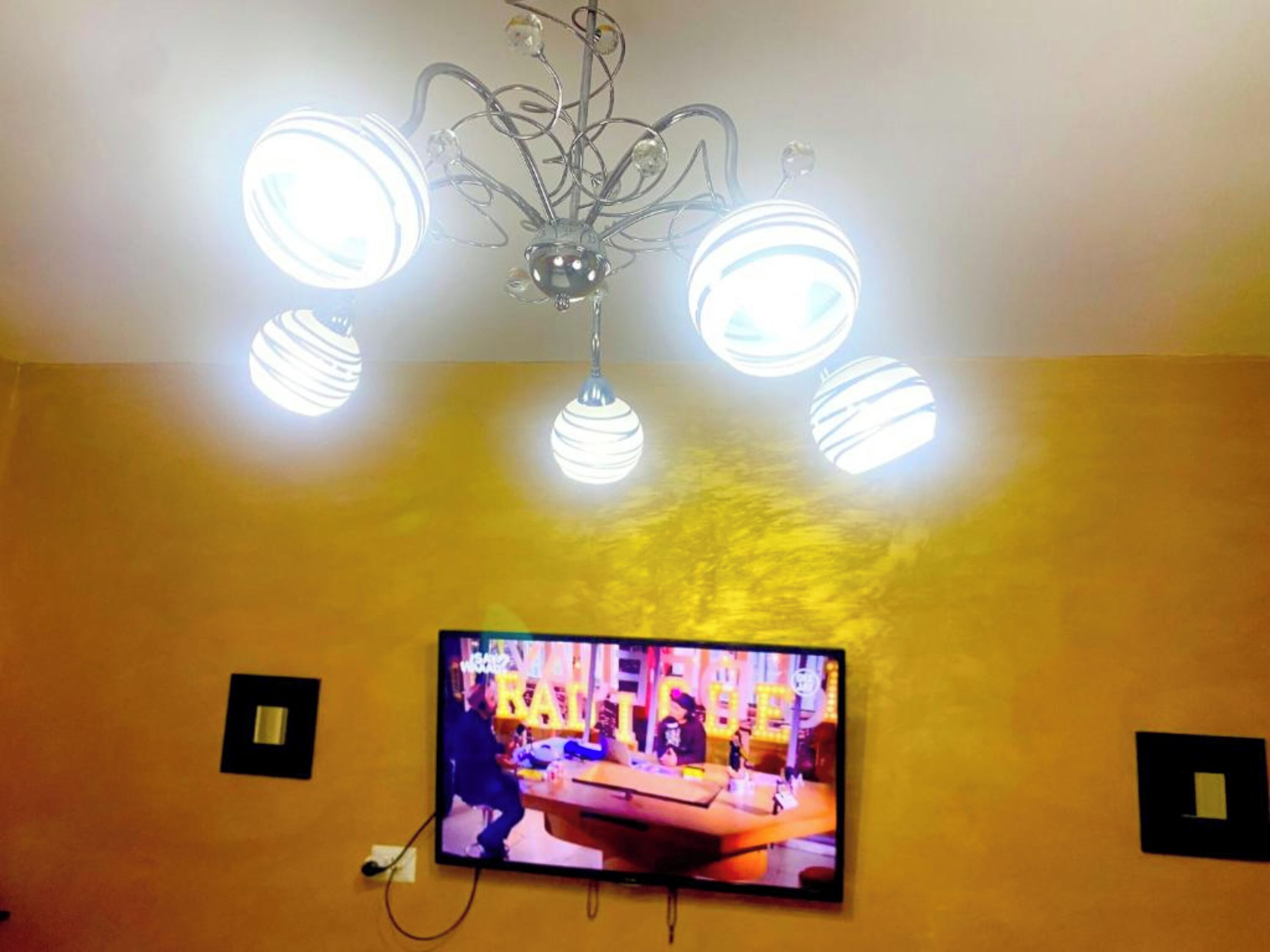 A television mounted on a yellow wall with a chandelier above it.