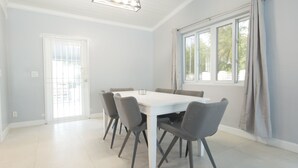 A dining area with a white table and grey chairs, large windows with curtains, and a door with a view of the outside.