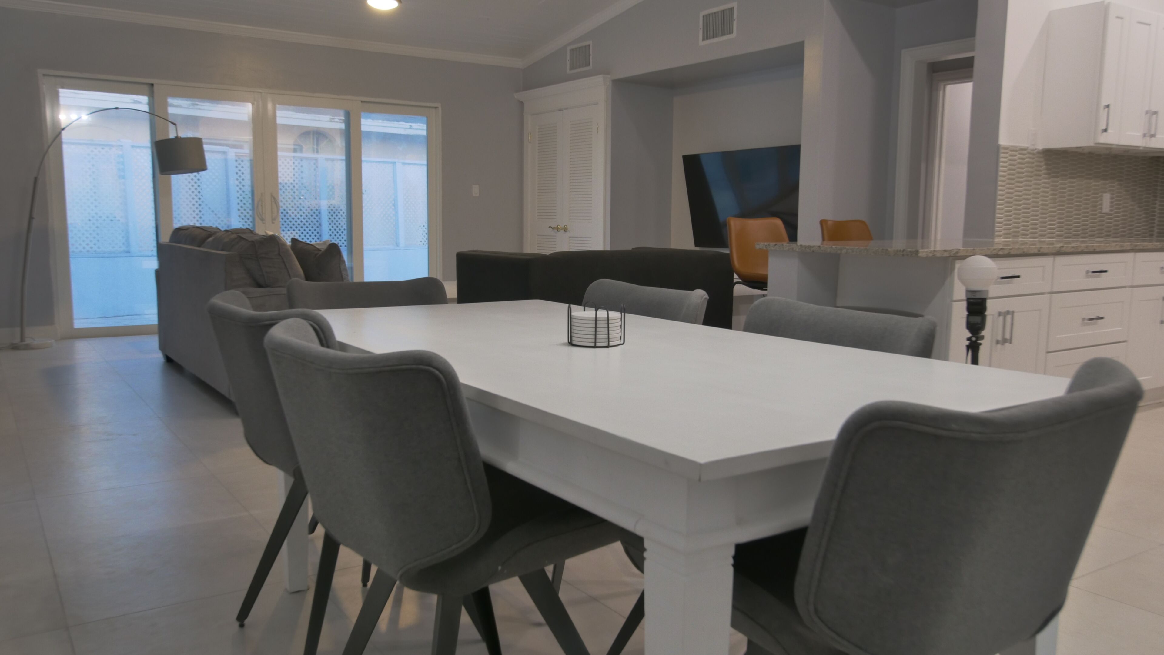 A modern kitchen with white cabinets, a dining area with a white table and grey chairs, and a grey sofa in the background.