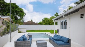 A backyard with a swimming pool, two grey wicker sofas with blue cushions, and a small table.