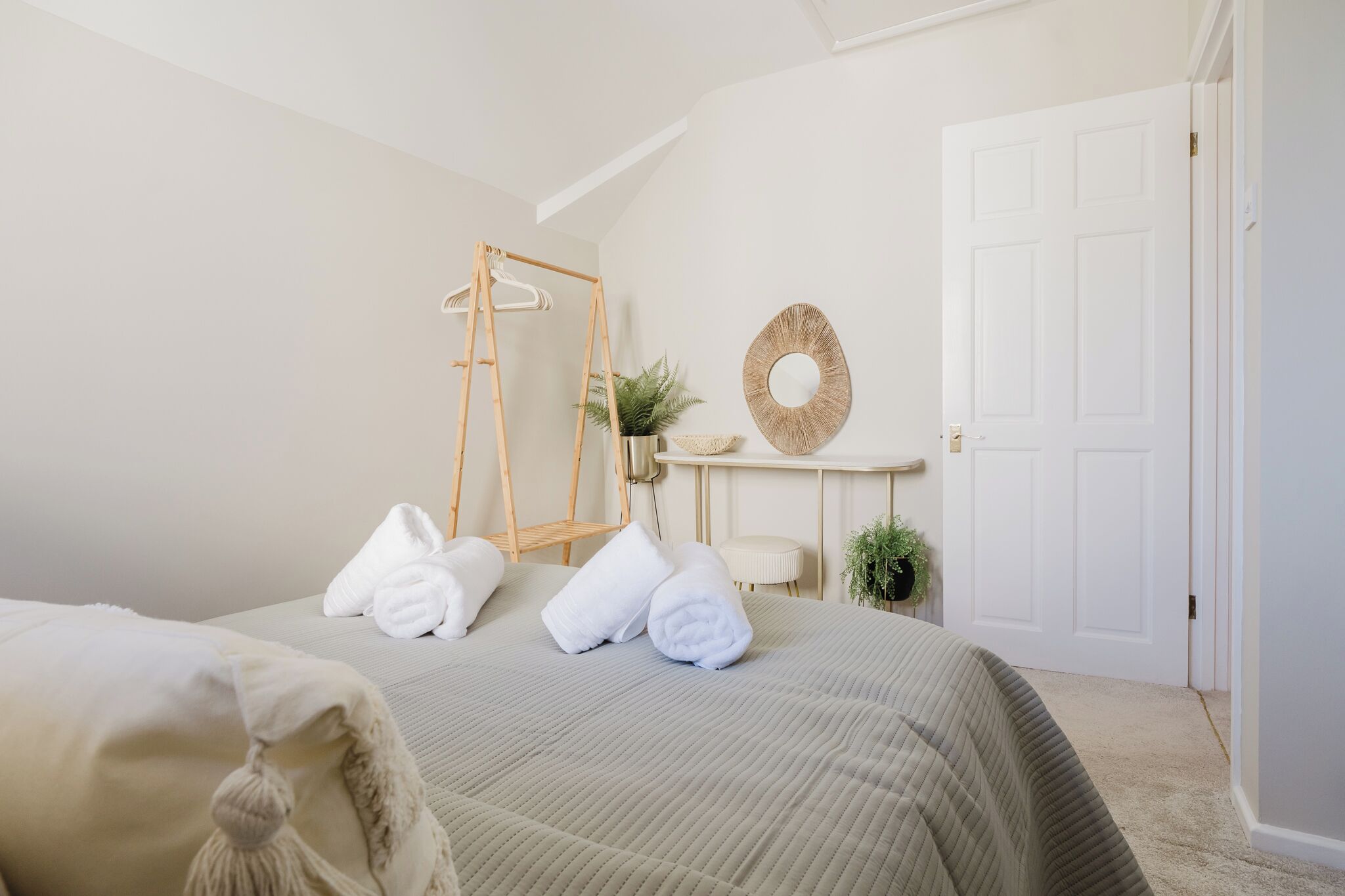 A neatly arranged bedroom with a bed, towels, a wooden ladder, a round mirror, and a door.