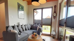 Een woonkamer met een grijze bank, een ronde rieten salontafel met fruit, een wandklok en een groot raam met jaloezieën.