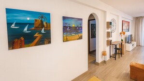A modern living room with a wooden floor, a dining area with a table and chairs, and two paintings on the wall depicting coastal scenes. - Varanda do Sol – Private Terrace with Sea View in Funchal (Funchal)