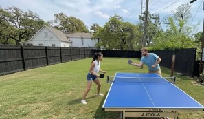 Deux personnes jouent au ping-pong dans un jardin, devant une table bleue, une clôture en bois et avec une maison en arrière-plan.