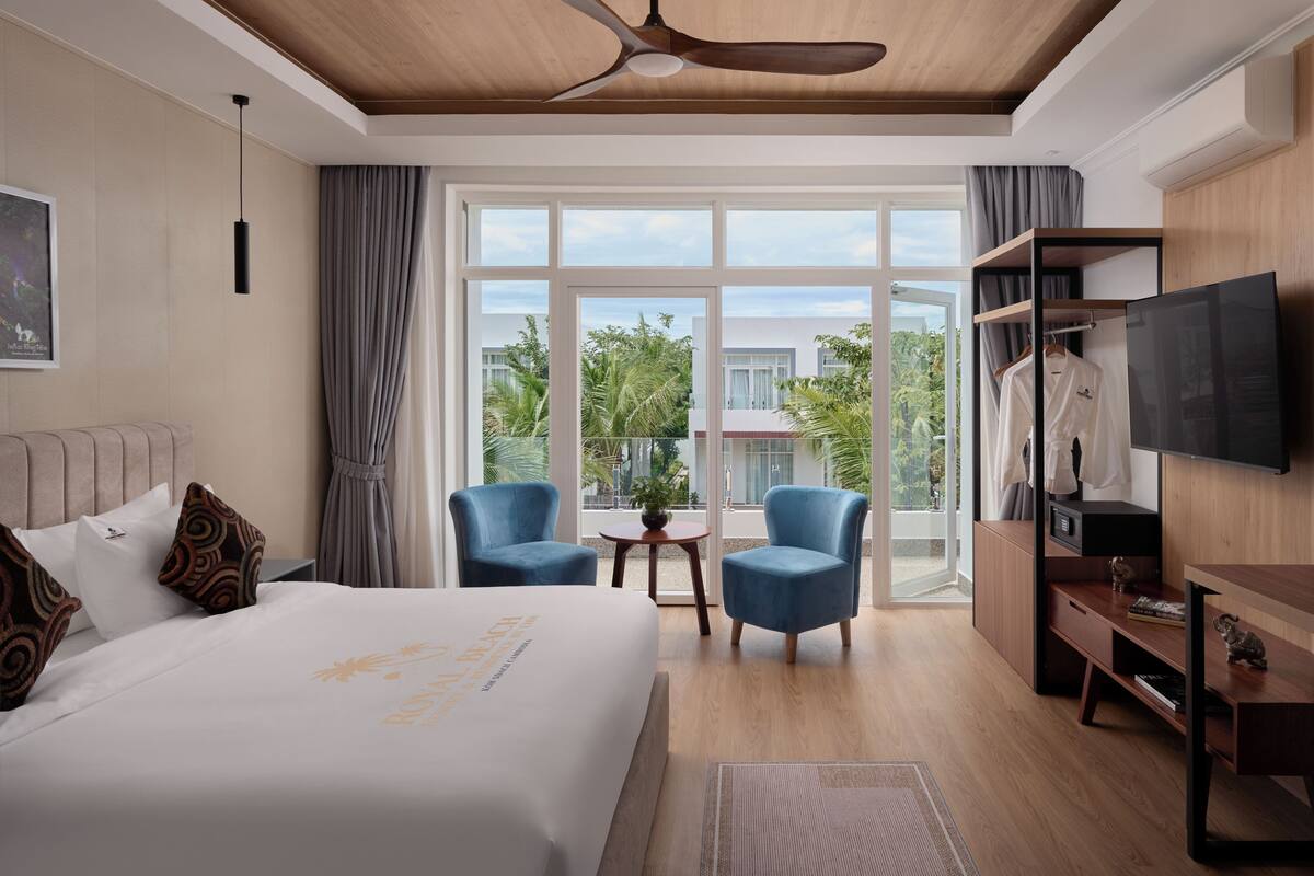 Habitación de hotel con una cama grande, dos sillones, televisor y balcón con vistas a edificios y árboles.