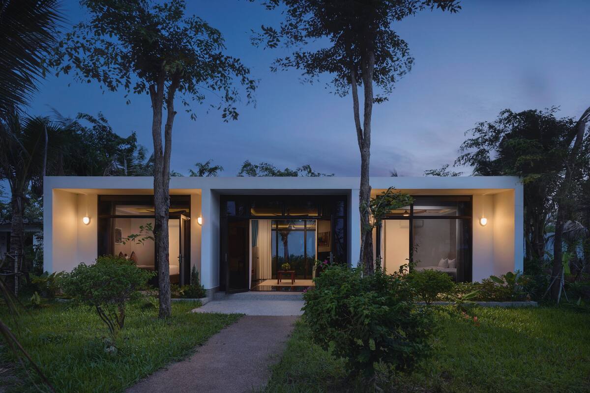 Una casa moderna de dos pisos con amplios ventanales y puertas de vidrio, rodeada de abundante vegetación.