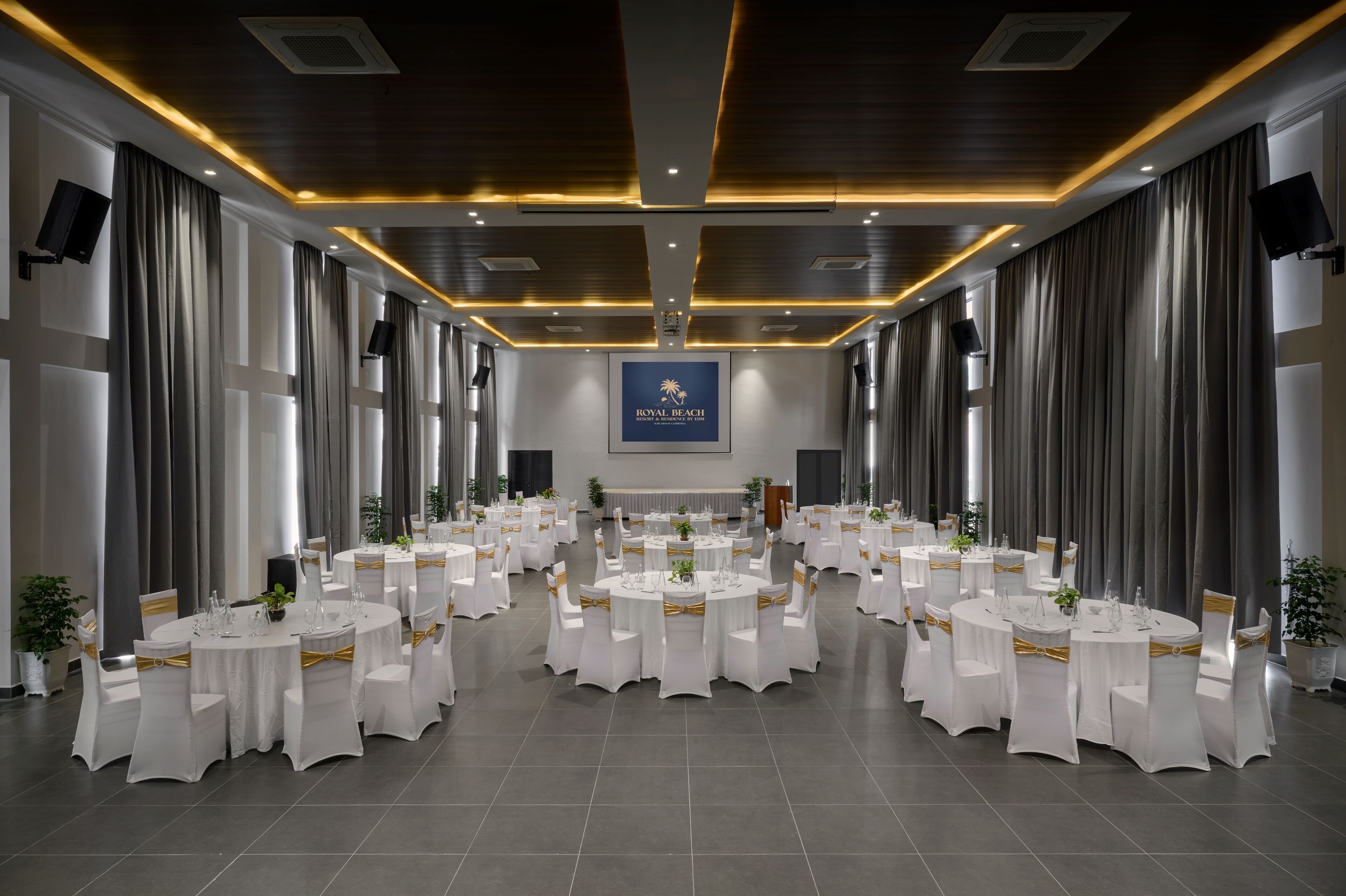A banquet hall with round tables, white chairs, and gold sashes, featuring a logo on the wall.