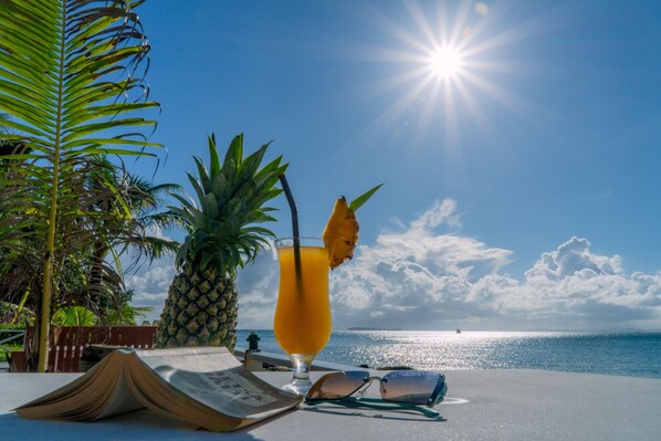 A tropical setting with a clear view of the ocean, a glass of orange juice with a straw, a pineapple, and a book on a table.