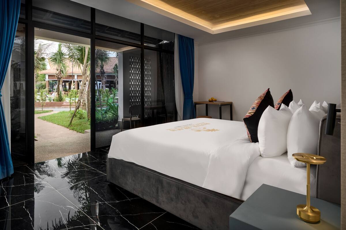 Habitación de hotel moderna con una cama grande, una mesita de noche y vistas a un patio con palmeras y una piscina.