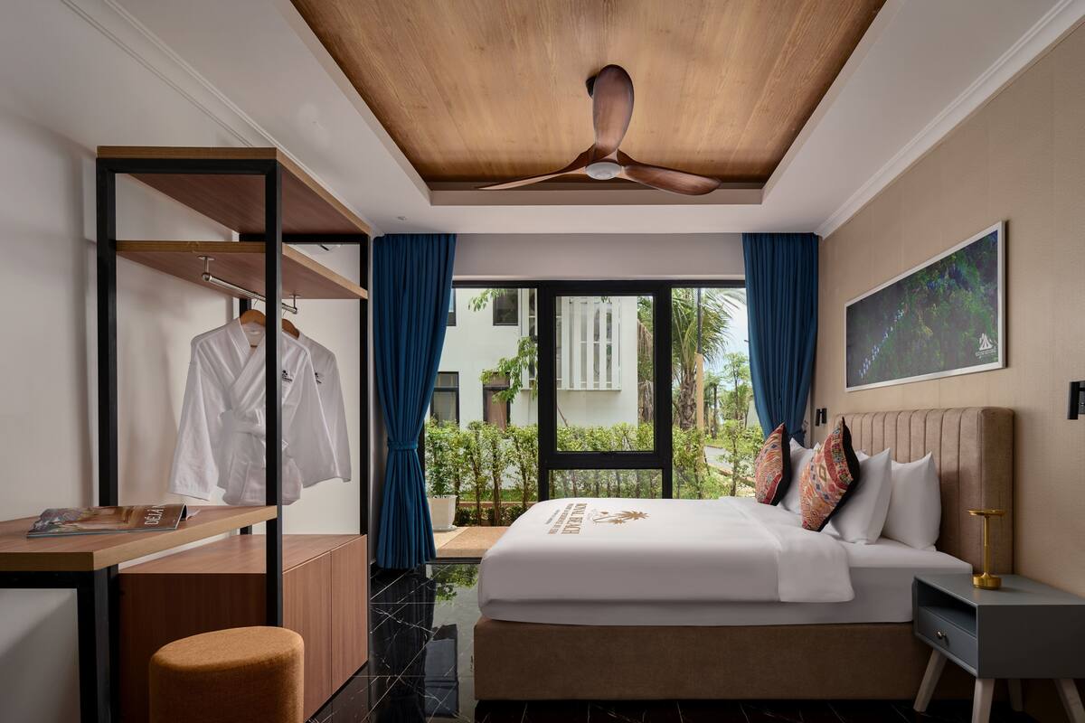 Habitación de hotel con cama, escritorio, armario, ventilador de techo y ventana con vistas a zonas verdes.