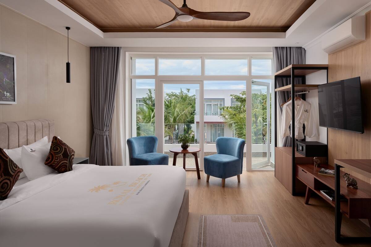 Una habitación de hotel con una cama grande, dos sillones azules, una mesita pequeña, un ventilador de techo y un televisor sobre un soporte de madera.