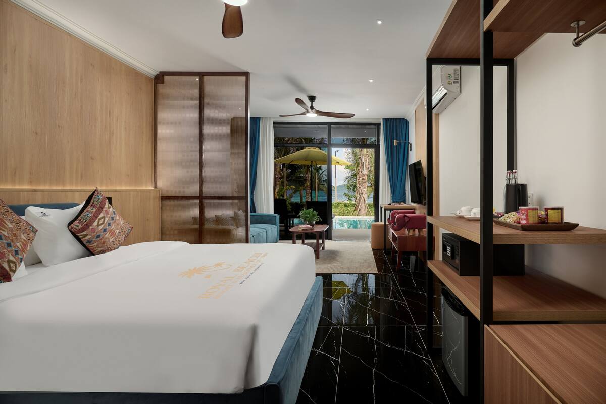 Una habitación de hotel moderna con una cama grande, un ventilador de techo, vista a la piscina y un comedor con mesa y sillas.