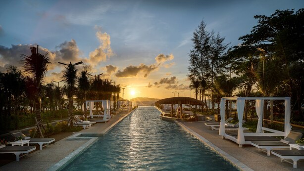 Una piscina con sillas de descanso y sombrillas, rodeada de árboles y un atardecer de fondo.