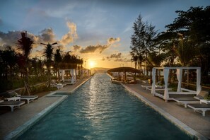A pool with lounge chairs and umbrellas, surrounded by trees and a sunset in the background.