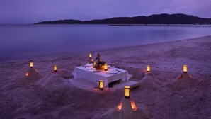 Montículos de arena rodean un espacio para comer frente al mar, con una mesa y sillas y faroles.