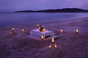 Ein Essbereich am Strand mit Tisch und Stühlen, umgeben von Sandhügeln mit Laternen.