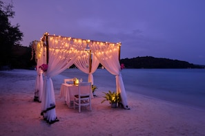 A beachside tent with draped fabric and string lights, a table with chairs, and potted plants.
