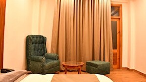 A room with a bed, a green armchair, a small round table, and a large window with curtains. - Mountain Majesty Manali (Manali)