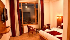 A hotel room with a bed, a TV mounted on the wall, a small table, and a window with curtains. - Mountain Majesty Manali (Manali)