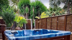 A hot tub with a wooden exterior, surrounded by a wooden fence and various tropical plants.