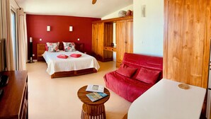 A bedroom with a bed, a red sofa, a wooden wardrobe, a ceiling fan, and a small round table with books.
