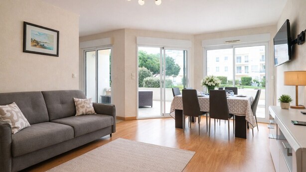 A modern living room with a grey sofa, a dining table with four chairs, and a TV mounted on the wall.
