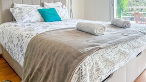 Ein ordentlich bezogenes Bett mit einer blumigen Tagesdecke, einem türkisfarbenen Kopfkissen und einem aufgerollten weißen Handtuch.