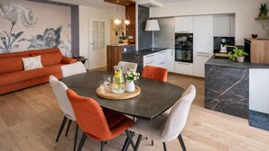 Eine moderne Küche mit einer zentralen Kochinsel, ein Essbereich mit dunklem Tisch und orangefarbenen Stühlen sowie eine Wohnbereich mit einer Couch und einer Wanddekoration.