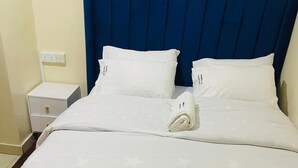 Una cama bien hecha con sábanas blancas y un cabecero azul, junto a una mesita de noche blanca y un enchufe.