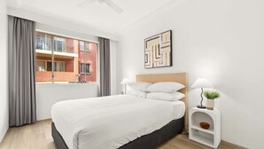 A modern hotel room with a large bed, a window with curtains, a headboard, a wall-mounted abstract art piece, and a bedside table with a lamp and a small plant.