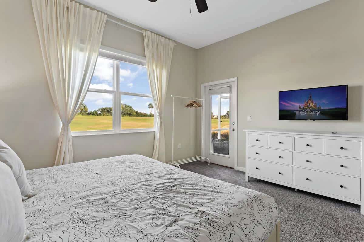 Un dormitorio con cama, televisor en la pared, cómoda y ventana con cortinas.