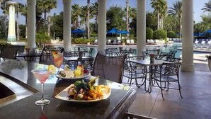 Un área de comedor al aire libre con mesas y sillas, una piscina al fondo y palmeras.