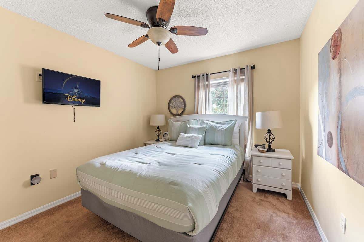 Une chambre à coucher avec un lit, des tables de chevet, un ventilateur de plafond, une télévision et un tableau au mur.